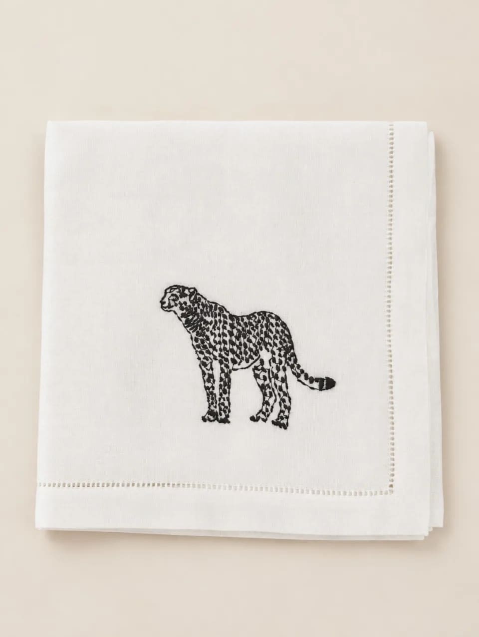 Servilleta de lino blanco con vainica y un leopardo negro bordado, doblada sobre un mantel de hojas